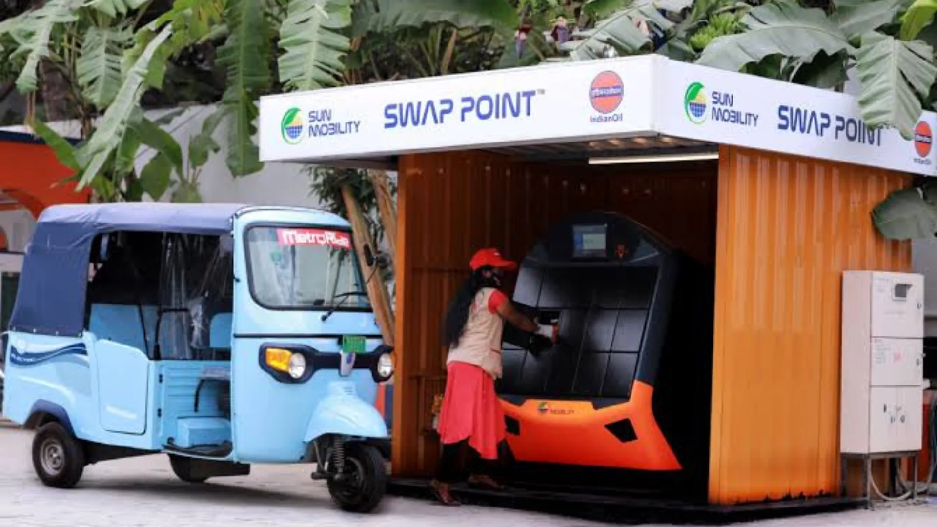A woman swaps a battery for a blue electric rickshaw at a SUN MOBILITY SWAP POINT kiosk in India, located at an IndianOil station.