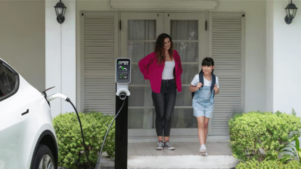 Mother and young daughter with a backpack standing near a white EV charging station mounted on a pedestal outside a residential garage door.