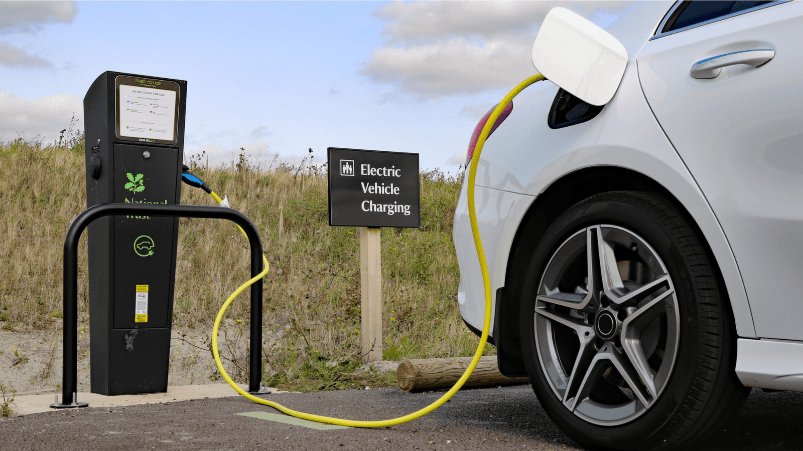 Electric Vehicle Charging at Public Station with Yellow Cable Connected to EV A white electric car charging at a public EV station with a yellow charging cable and a sign reading “Electric Vehicle Charging” beside the charger.